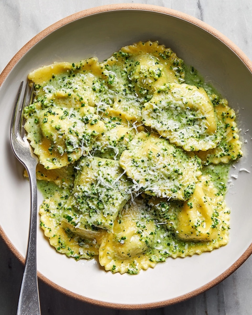 A white bowl holds a serving of ravioli covered in a thick green pesto sauce with visible herb specks. The ravioli pieces are yellow with a slightly glossy texture and scalloped edges. The pasta is layered with some pieces overlapping, and the pesto sauce evenly coats the ravioli, pooling lightly at the bottom. Finely grated white cheese is sprinkled on top, adding a delicate texture contrast. A silver fork rests inside the bowl on the left side. The bowl sits on a white marbled surface. Photo taken with an iphone --ar 4:5 --v 7