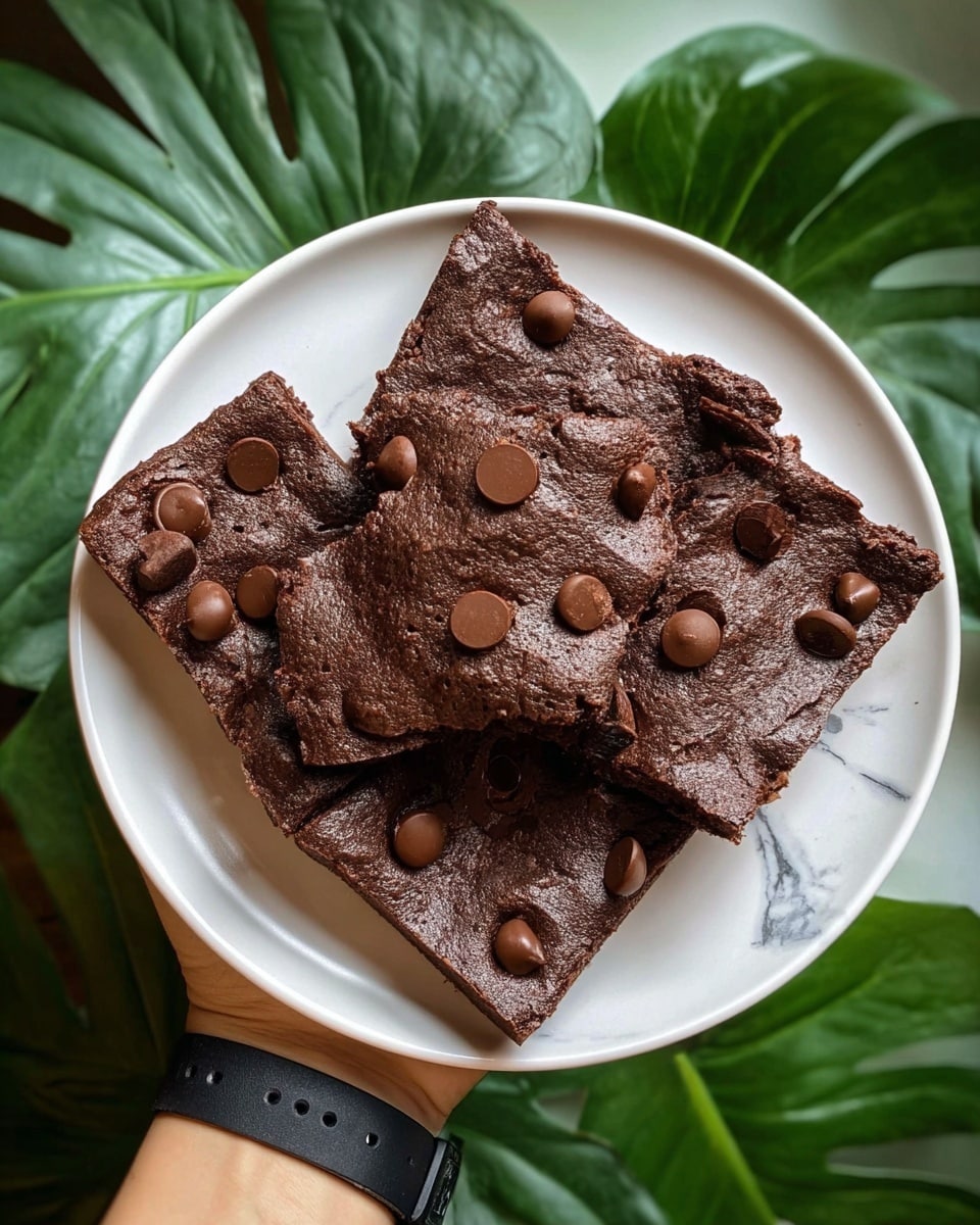 A white round plate holds five rich, dark brown chocolate brownies, each topped with several round chocolate chips. The brownies have a slightly wrinkled texture on top, showing gooey moistness, with some edges slightly raised and curved. The pieces are arranged in a loose pile, with some overlapping, and show a dense, fudgy texture. A woman's hand, wearing a black wristband, supports the plate, which is set against a background of large green leaves on a white marbled surface. photo taken with an iphone --ar 4:5 --v 7