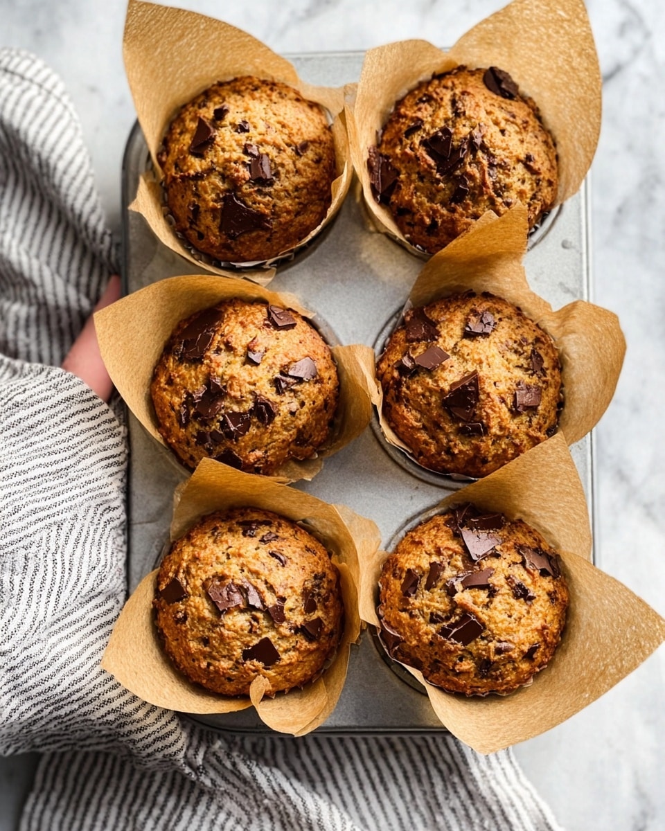 The image shows six golden brown muffins in a silver metal muffin tray, each wrapped in light brown parchment paper. The muffins have a rough texture and are topped with chunks of melted dark chocolate scattered unevenly across the surface. The tray rests on a white marbled surface, with a striped cloth napkin visible at the bottom left corner. A woman's hand is gently holding the edge of the tray from the left side. The overall lighting is soft and natural, highlighting the warm tones of the muffins. photo taken with an iphone --ar 4:5 --v 7