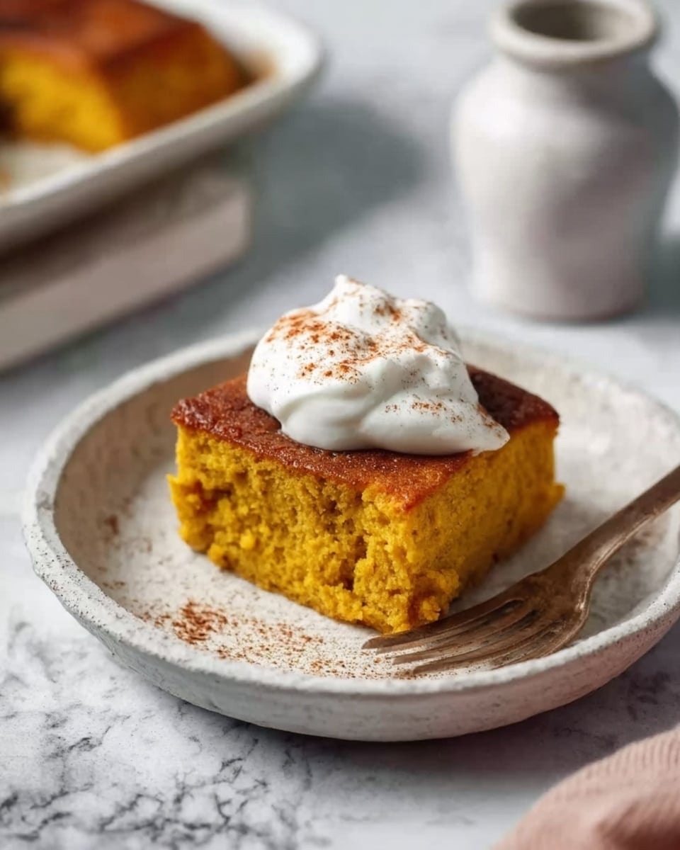 A square piece of golden yellow pumpkin cake sits on a white ceramic plate with a slight texture. The cake has a soft, moist texture with a light brown caramelized top layer. On top of the cake, there is a dollop of white cream sprinkled lightly with cinnamon powder. Next to the cake on the plate is a small silver fork resting on the edge. The plate is set on a white marbled surface with a small white ceramic jug in the background. Photo taken with an iphone --ar 4:5 --v 7