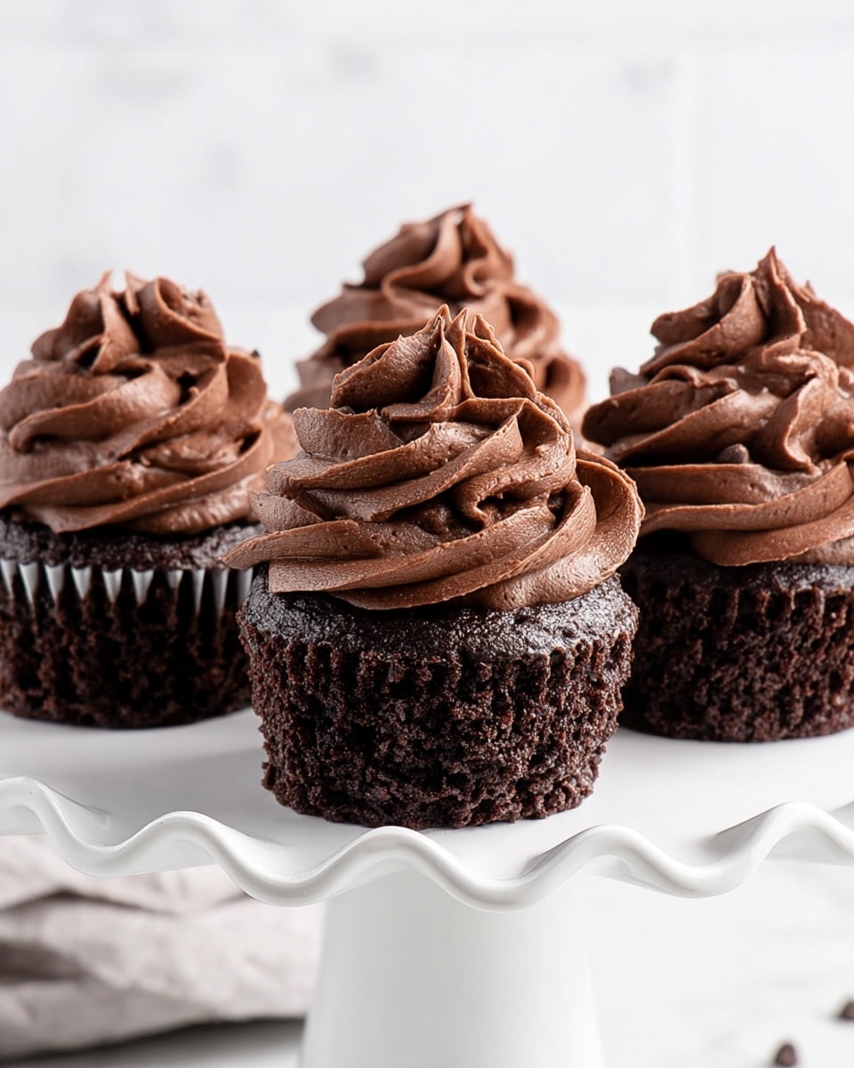 The image shows four rich chocolate cupcakes placed on a white ceramic cake stand with wavy edges, set against a white marbled surface. Each cupcake has a dark, moist chocolate base with visible chocolate chips embedded in the cake. On top of each base is a thick, swirled layer of smooth, dark chocolate frosting that has a rich, velvety texture and elegant, wavy folds. The frosting extends upwards in a high, decorative peak, covering the entire top surface of the cupcakes. The background is bright and softly blurred, emphasizing the dark, glossy cupcakes in the center of the image. Photo taken with an iphone --ar 4:5 --v 7