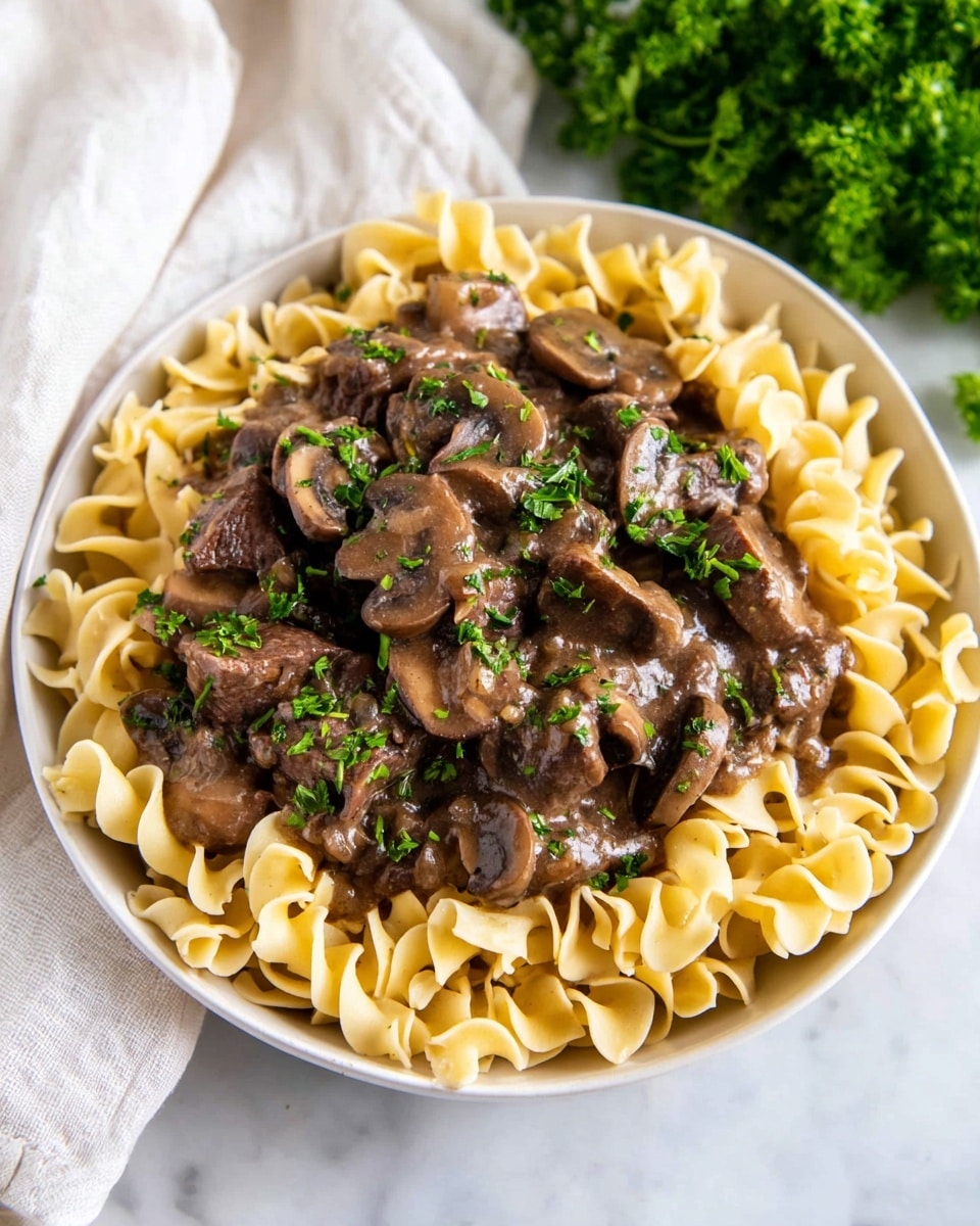 A white bowl filled with three layers: the bottom layer has light yellow, curly egg noodles arranged in a thick ring near the edge. The middle layer is a dark brown creamy sauce containing sliced mushrooms and tender pieces of beef. The top layer is sprinkled with bright green chopped parsley, adding fresh color. The dish is placed on a white marbled surface with a white cloth and green parsley in the background. photo taken with an iphone --ar 4:5 --v 7