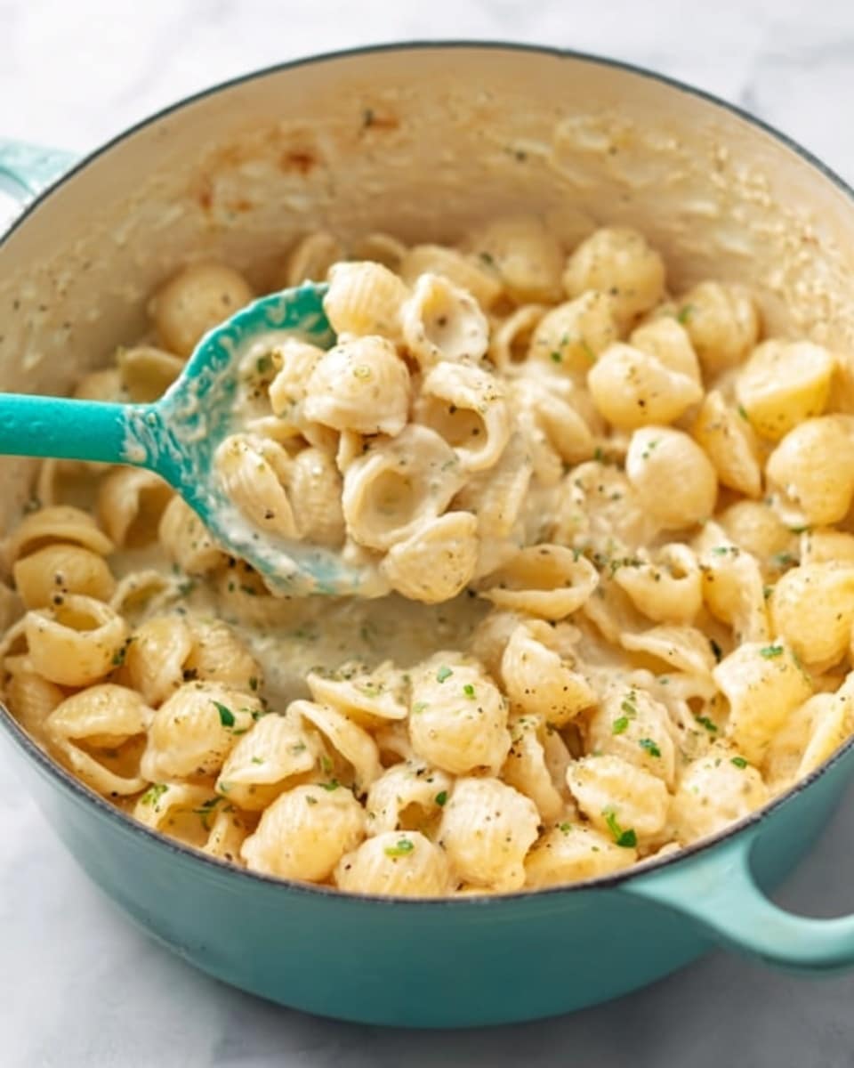 The dish shows small round pasta shells covered in a creamy white sauce with a few small green herb specks mixed throughout. The pasta is inside a white pot, and a turquoise spoon is lifted from the pot’s right side, holding some of the pasta. The sauce looks smooth and thick, making the pasta glisten. The scene is bright with a white marbled surface underneath the pot. photo taken with an iphone --ar 4:5 --v 7