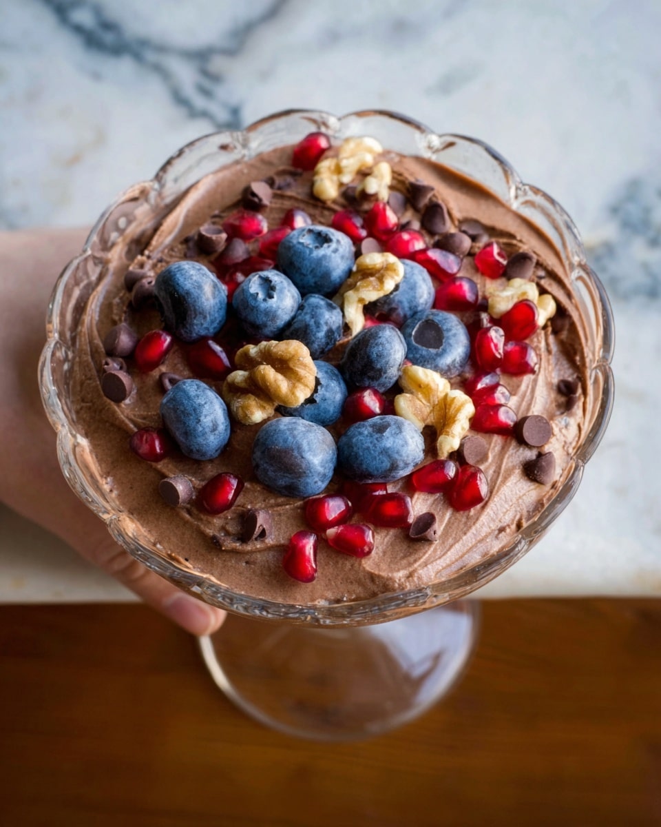 A white bowl filled with smooth chocolate mousse sits in the center, topped with fresh blueberries, red pomegranate seeds, chocolate chips, and small crumbly pieces. Around the bowl on a white marbled surface, there are sections of various foods arranged in a circular pattern: bright blue blueberries, crunchy light brown granola, fresh red strawberries with green tops, small chunks of light brown peanut butter cups, white shredded coconut, dark brown chocolate chips, and shiny red pomegranate seeds. The mix of colors and textures creates a rich, inviting look. photo taken with an iphone --ar 4:5 --v 7