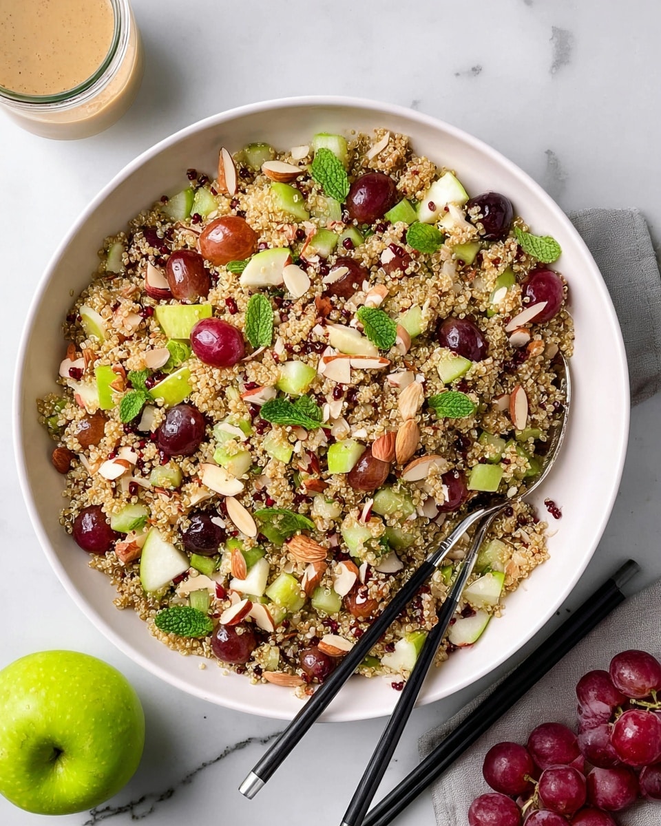 A large white bowl filled with a quinoa salad that has a mix of small beige quinoa grains, halved red grapes, chopped green and red apple pieces, and thin slices of celery. The salad is topped with small almond slivers and fresh green mint leaves scattered on top. A silver spoon with a black handle rests in the bowl, with black chopsticks leaning on the bowl's edge. The bowl is placed on a white marbled surface with a green apple, a small bunch of red grapes, and a glass jar with a light beige dressing partially visible nearby. Photo taken with an iphone --ar 4:5 --v 7