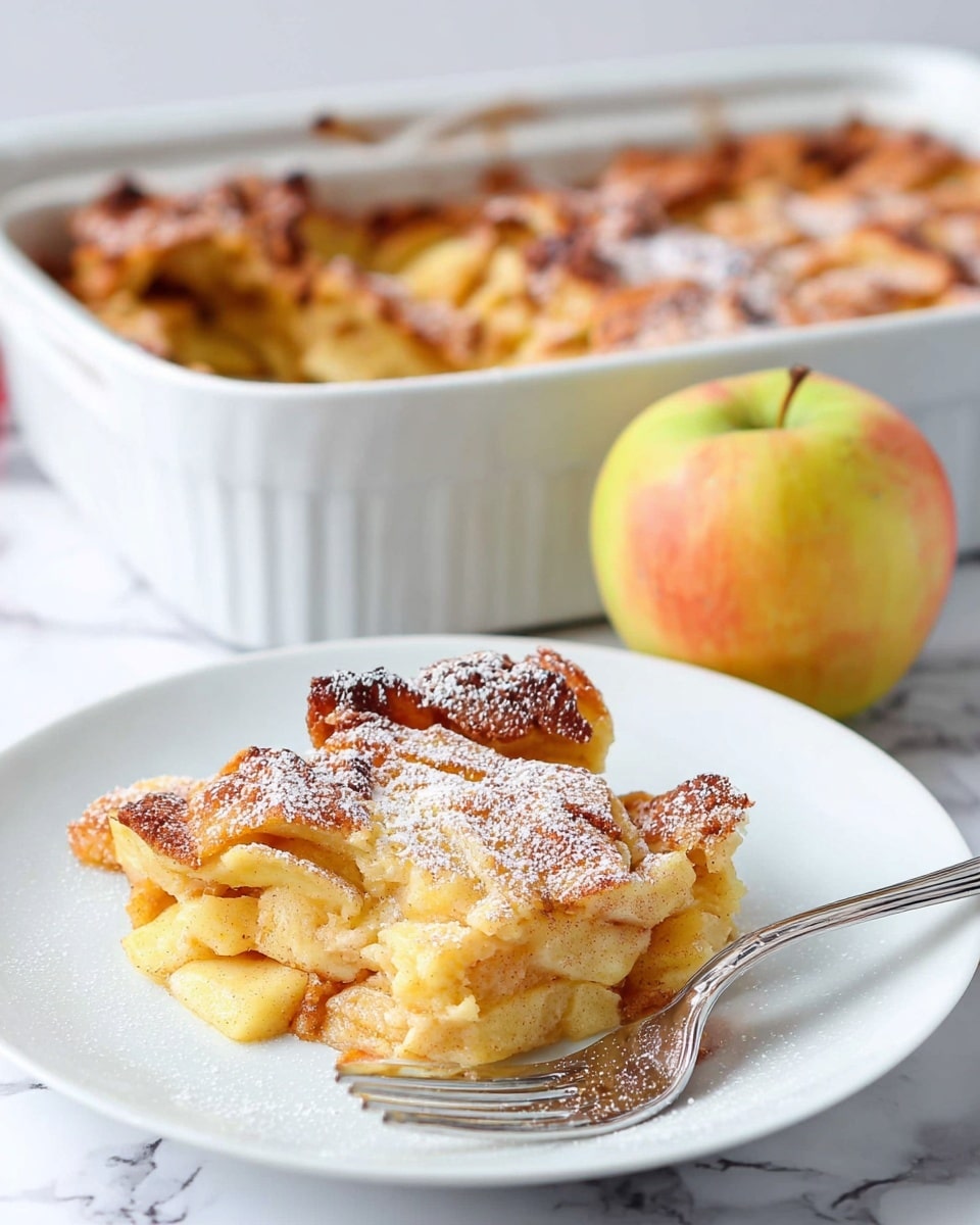 A white plate holds a single serving of a golden-brown baked apple dish, with a slightly crispy, uneven top layer dusted with powdered sugar, showing soft, pale yellow inside layers mixed with tender apple pieces. Behind the plate is a white baking dish, filled with more of the same baked apple dessert with a browned, rough surface. Next to the plate is a fresh apple with yellow and red skin, and a silver fork rests on the plate. The setting is on a white marbled surface. Photo taken with an iphone --ar 4:5 --v 7