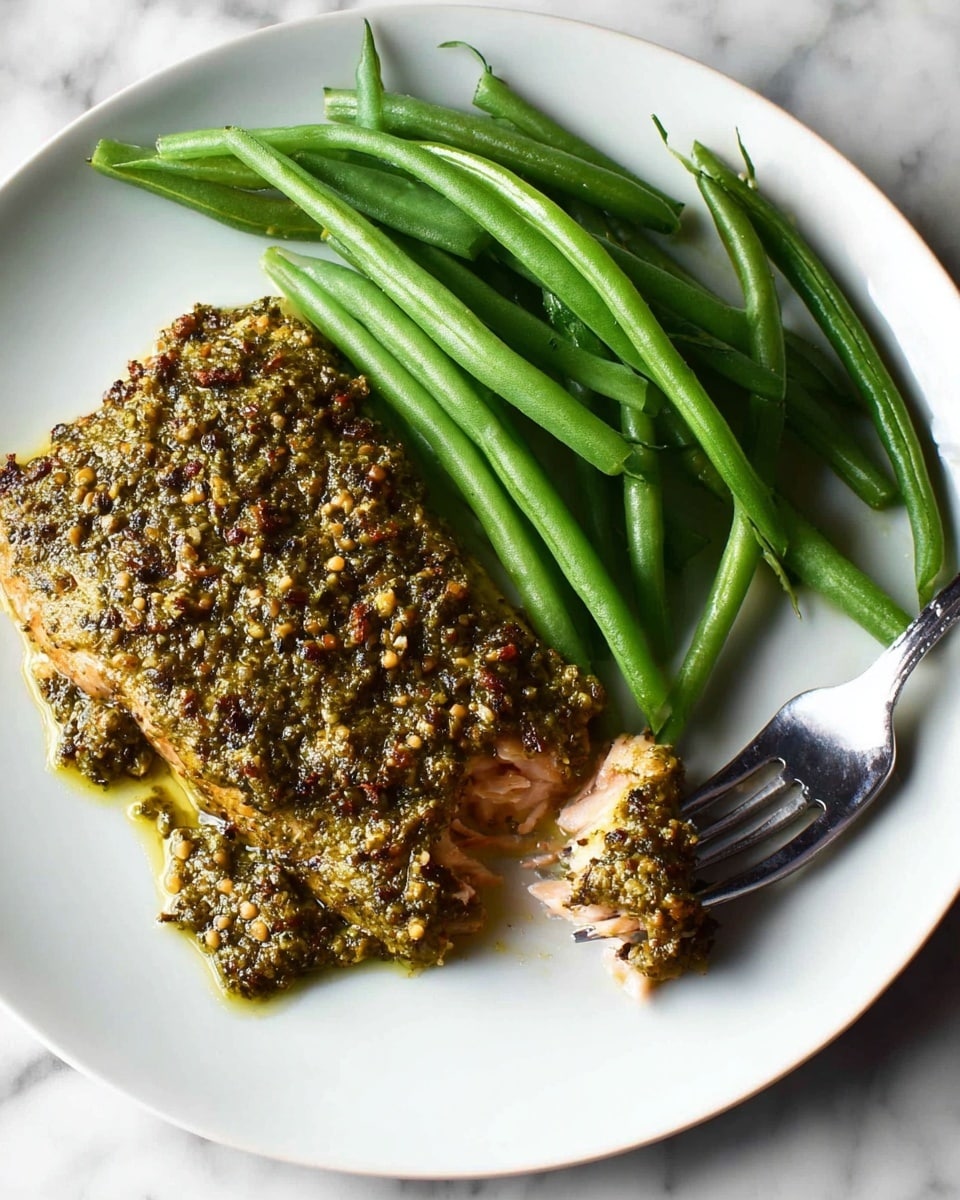 A white plate on a white marbled surface holds a serving of fish topped with a thick greenish brown nutty pesto crust, displaying a rough and textured layer with small chunks. Below the crust, tender pink fish flesh is slightly visible breaking apart at the edges. Next to it, a neat pile of long, fresh green beans with a smooth texture and slight shine lies close to a silver fork positioned at the plate's edge. The simple, clean presentation highlights the natural colors and textures of the food. photo taken with an iphone --ar 4:5 --v 7