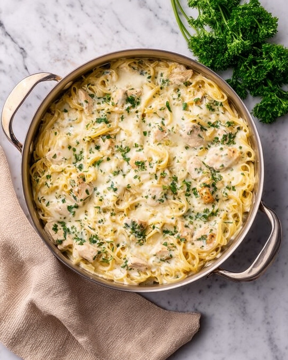 The image shows a large metal pan filled with a creamy pasta dish. The base layer is creamy white cheese sauce mixed with cooked chicken pieces, followed by tender pasta strands that are coated in the sauce. There are green herb pieces sprinkled evenly throughout, giving a touch of color and freshness. The top layer is melted cheese, slightly browned and bubbly, with larger green herb flakes scattered over it. The pan is set on a white marbled surface, next to a beige cloth napkin and a green parsley sprig for decoration. Photo taken with an iphone --ar 4:5 --v 7