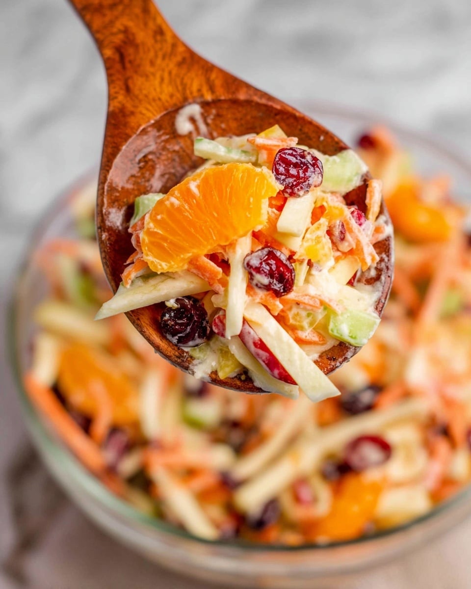 A clear glass bowl filled with a colorful salad showing multiple layers and textures; the base layer consists of thin, long strips of light green celery and white apple, mixed with bright orange mandarin segments scattered evenly throughout; thin, vibrant orange carrot sticks are layered across the salad creating a rough texture, interspersed with dark red dried cranberries adding color contrast; the salad is coated lightly with a creamy dressing that gives a slight shine to the ingredients; the bowl is placed on a soft gray cloth with a herringbone pattern which rests on a white marbled surface; next to the bowl, two wooden salad spoons with smooth textures are arranged crossing each other; photo taken with an iphone --ar 4:5 --v 7
