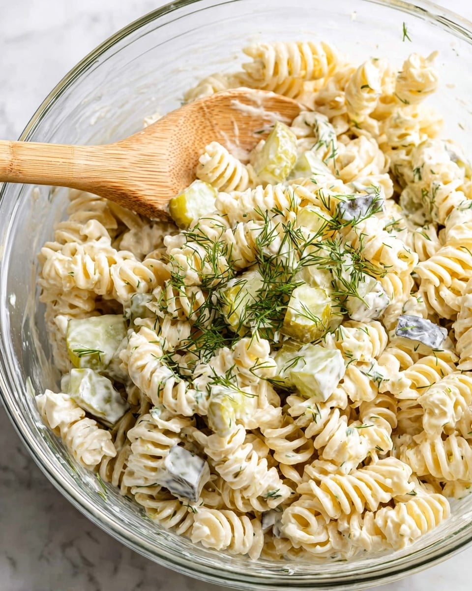 A clear glass bowl filled with a creamy pasta salad, featuring three main layers mixed together: short, yellow rotini pasta as the base, diced pale green celery pieces, and bits of light yellow potato, all coated in a thick white creamy dressing. The salad is sprinkled with fine green dill herb over the top for a fresh touch. The bowl sits on a white marbled textured surface with soft natural light highlighting the different textures of the pasta and vegetables. Photo taken with an iphone --ar 4:5 --v 7