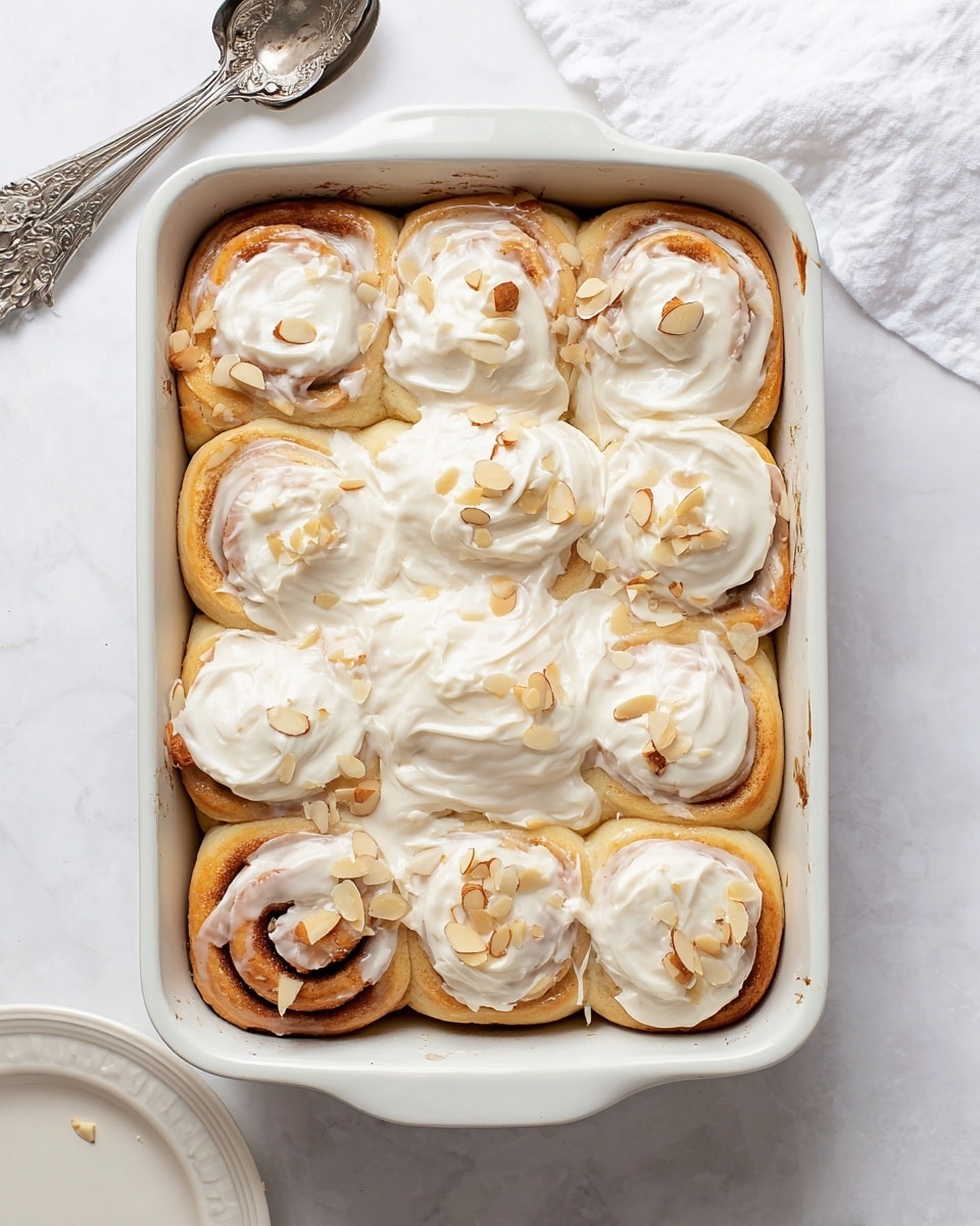 A white rectangular baking dish holds eight cinnamon rolls arranged in two rows of four, each roll thickly coated with a shiny white icing that drips slightly down the sides. On top of the icing are thin, pale almond slices scattered unevenly on each roll, dusted further with fine white powdered sugar. The cinnamon rolls have a light golden-brown dough visible beneath the icing on some edges. A woman's hand with a cream-colored knitted sweater and pink nails reaches toward the top right roll. The dish rests on a white marbled surface with a soft cream-colored textured cloth and a small bowl of almond slices partially visible nearby. Photo taken with an iphone --ar 4:5 --v 7