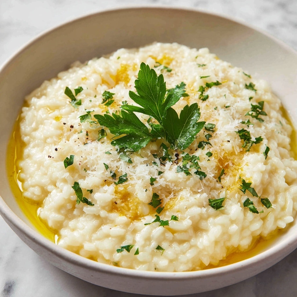 A close-up of a shallow pan filled with creamy yellow risotto with visible plump rice grains, topped with thin white parmesan cheese shavings and small green sprigs of herbs, with a few black pepper specks sprinkled on top. A shiny gold spoon rests inside the pan on the left side. The background shows a soft white marbled texture. photo taken with an iphone --ar 4:5 --v 7