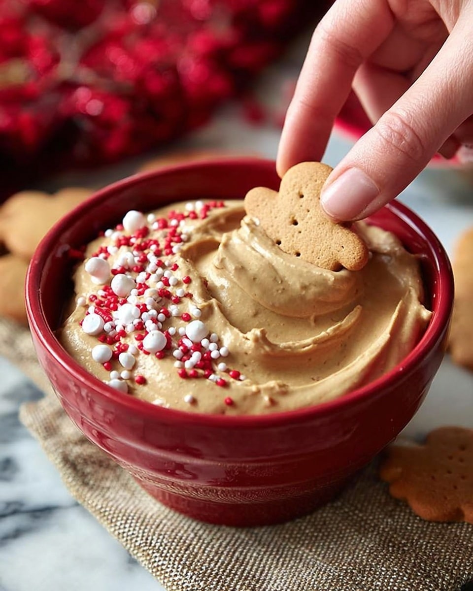 A red bowl filled with a thick, smooth, light brown creamy dip, swirled on top with soft peaks. The dip is decorated with small red and white round sprinkles and tiny gingerbread man-shaped brown and white sprinkles scattered over the top. A woman's hand is dipping a small round beige cookie into the creamy dip. The bowl is set on a textured beige cloth with hints of red fabric in the background, all on a white marbled surface. photo taken with an iphone --ar 4:5 --v 7