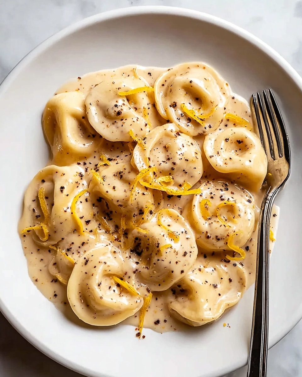 The image shows a white plate filled with several pieces of tortellini pasta covered in a creamy beige sauce. The tortellini have a smooth texture with slightly curved edges, and the sauce coats each piece evenly. Small red chili flakes are sprinkled across the top, adding tiny specks of deep red color. There are also bright yellow lemon zest shreds scattered on the tortellini, giving a fresh contrast to the creamy sauce. The background is a white marbled surface. photo taken with an iphone --ar 4:5 --v 7
