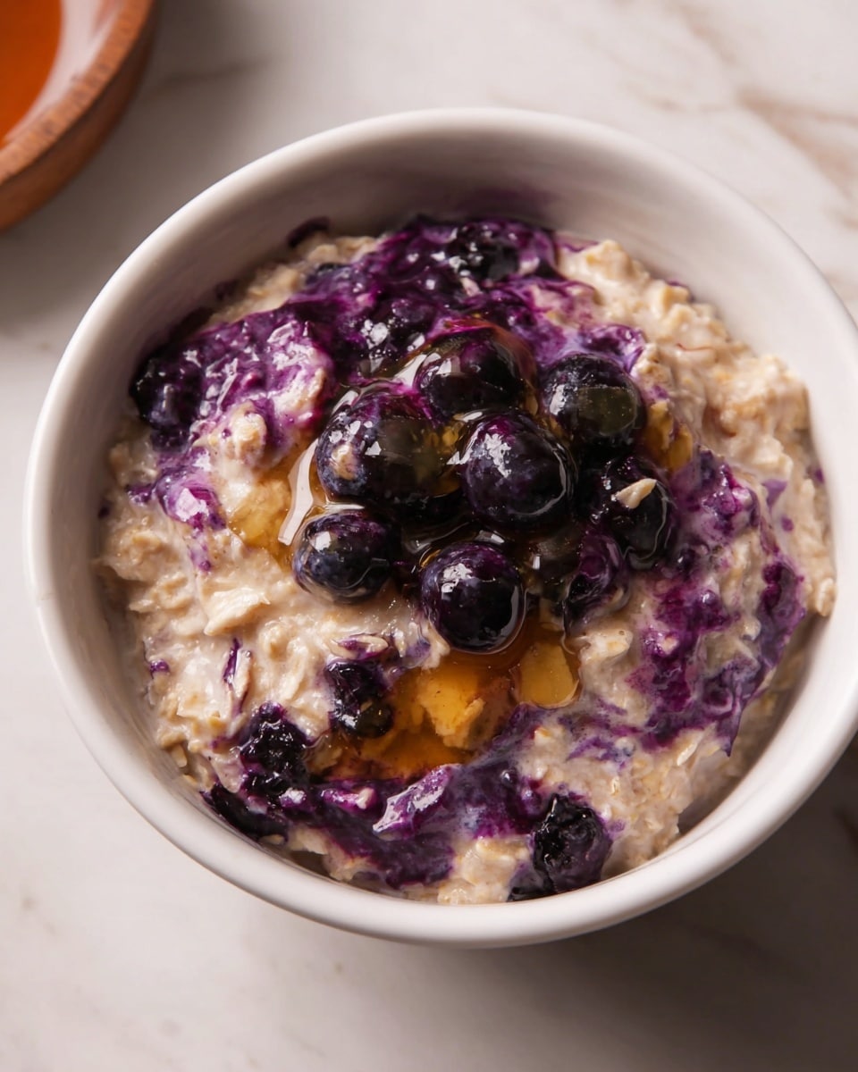 A white bowl filled with creamy oatmeal swirled with purple blueberry sauce and whole blueberries mixed inside. The oatmeal has a soft, thick texture with a light beige color, and the dark purple is spread unevenly across the top and bottom, creating a marbled effect. On top, a drizzle of golden syrup is falling onto the dish, adding a shiny, sticky layer. The bowl is on a white marbled surface, making the colors of the oatmeal and blueberries pop. photo taken with an iphone --ar 4:5 --v 7