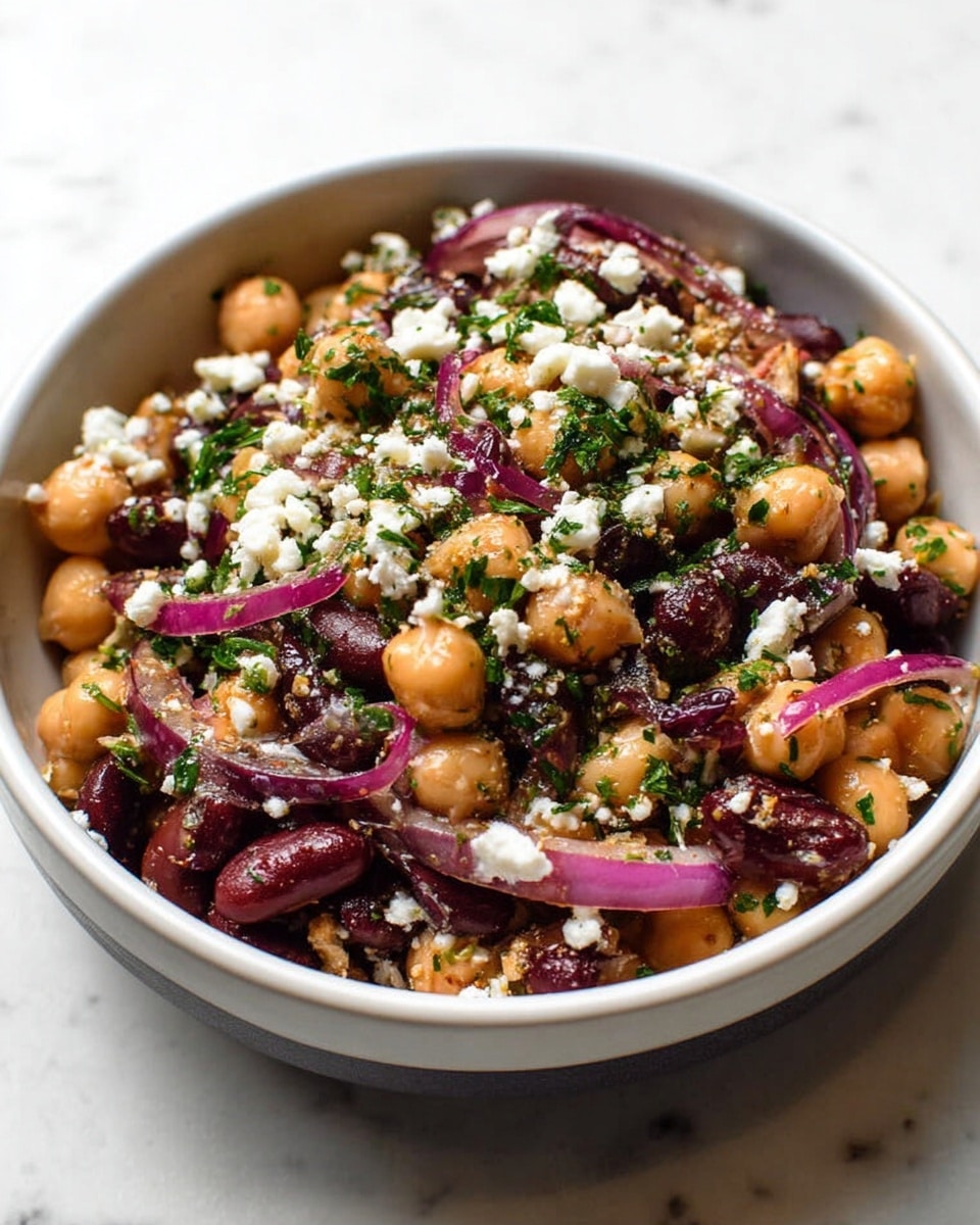 In a white bowl filled with a mixture of beige chickpeas and dark red kidney beans, there are thin slices of purple-red onion scattered throughout. The dish is topped with white crumbled cheese and finely chopped green herbs, creating a fresh and colorful look. The textures range from soft beans to crunchy onions and crumbly cheese. The bowl sits on a white marbled surface. photo taken with an iphone --ar 4:5 --v 7