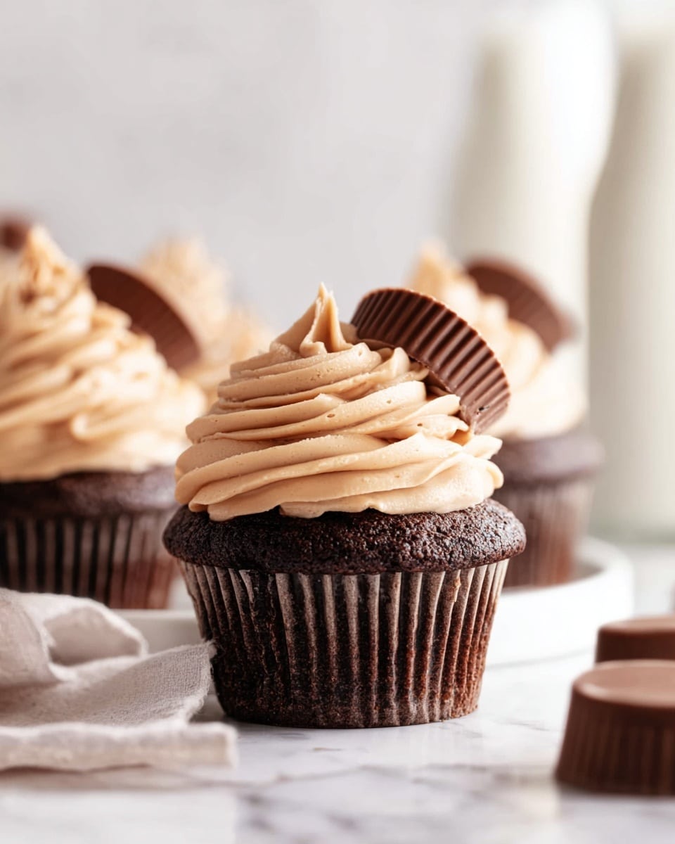 The image shows two dark chocolate cupcakes cut in half, revealing a gooey caramel center inside the rich, moist chocolate cake layer. On top of each cupcake is a thick layer of light brown, fluffy frosting with a creamy texture. Each cupcake is topped with a half of a smooth, shiny, dark chocolate peanut butter cup. The cupcakes rest on white cupcake liners, placed on a white marbled surface. In the background, there is a whole chocolate peanut butter cup near the cupcakes. photo taken with an iphone --ar 4:5 --v 7