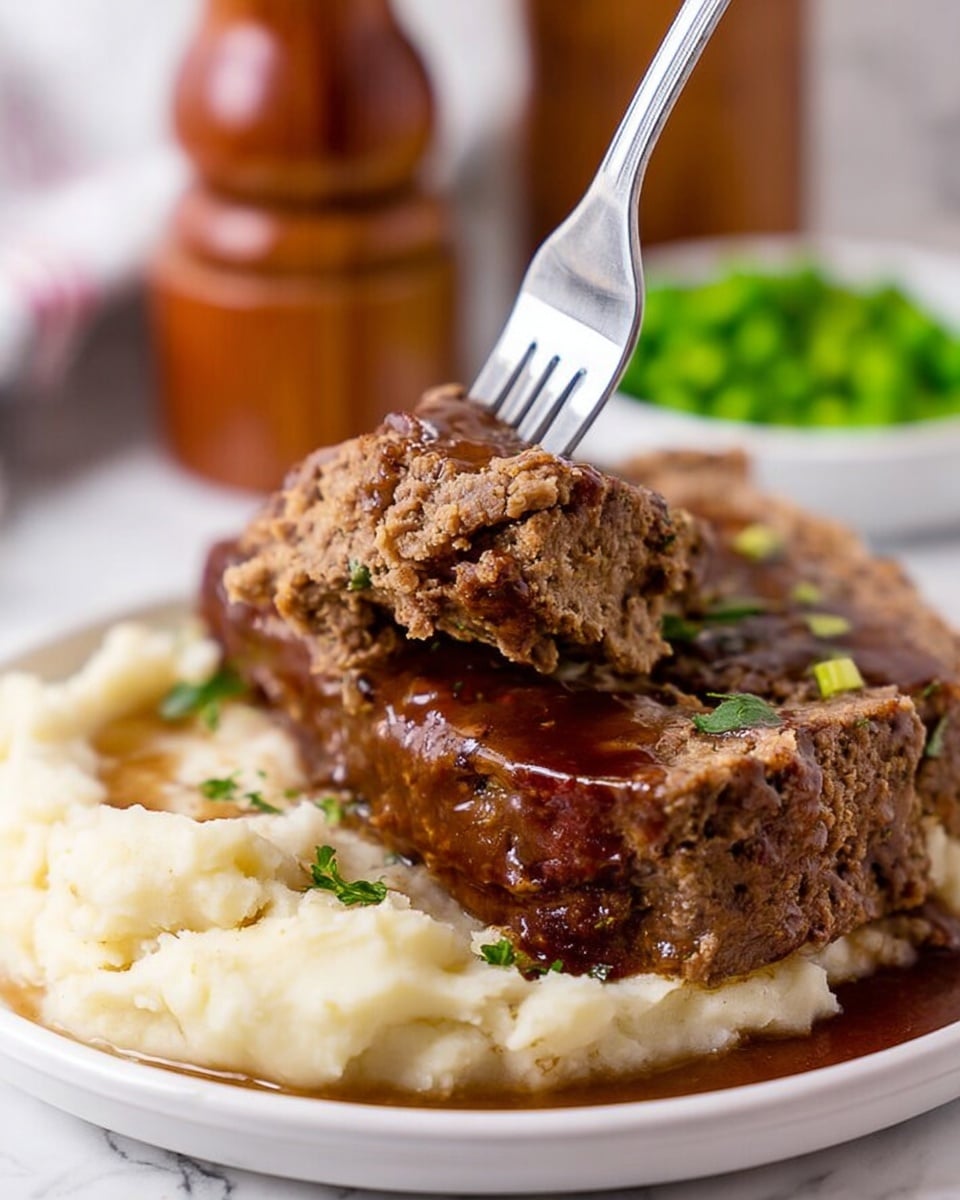 The image shows a close-up of a dish with three main layers on a white plate placed on a white marbled surface. The bottom layer is a bed of creamy, mashed potatoes with a smooth texture and a few green herb pieces sprinkled on top. Above the mashed potatoes lies a thick, brown meatloaf with a slightly rough surface, sliced into thick portions. The meatloaf is covered with a glossy, dark brown gravy that drips down onto the mashed potatoes. A fork is held close to the camera, lifting a chunk of the meatloaf, showing its crumbly and moist texture. In the background, there is a blurred wooden pepper grinder and some green peas. Photo taken with an iphone --ar 4:5 --v 7