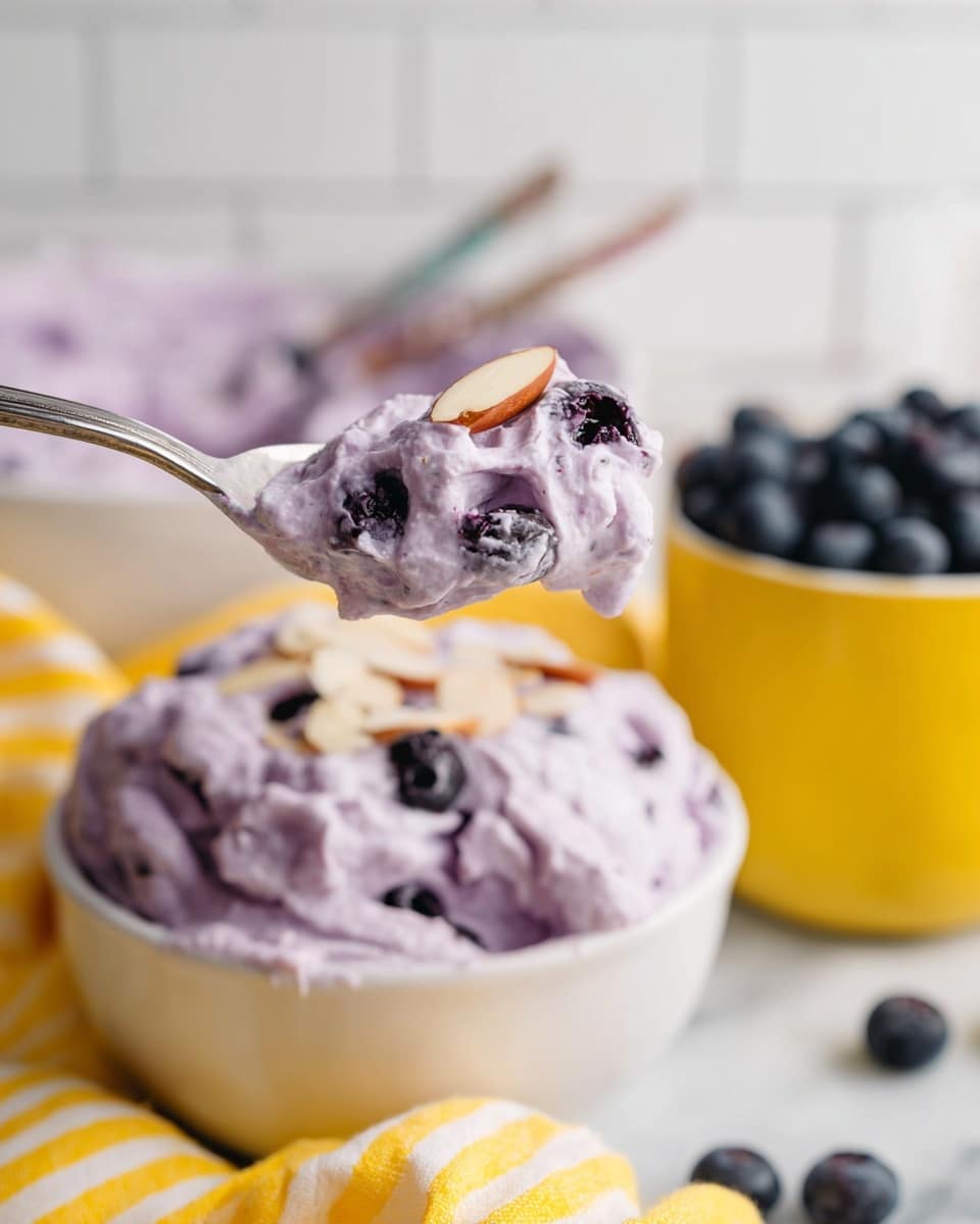 A large clear glass bowl filled with a thick, creamy, light purple mixture full of small darker purple bits, topped with a cluster of whole fresh blueberries in the center and scattered thin sliced almonds on the surface, surrounded by a few whole blueberries on a white marbled surface; in the background, another small clear glass container with the same purple mixture and a small white bowl filled with blueberries are visible, along with a yellow striped cloth. photo taken with an iphone --ar 4:5 --v 7