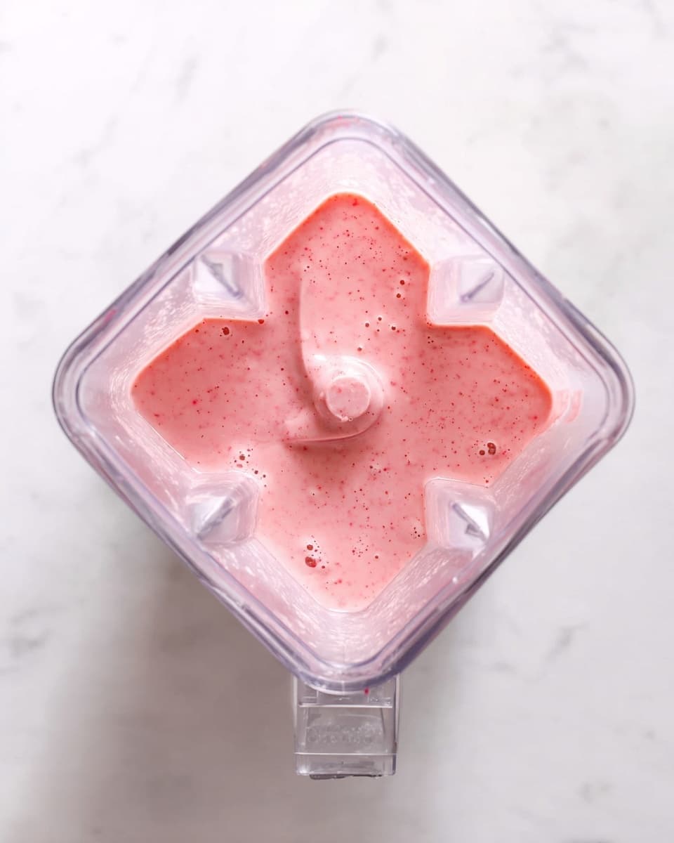 The image shows a blender filled with a smooth pink mixture that has a creamy texture and small bubbles on the surface, sitting on a white marbled texture. The blender jar is clear and square-shaped, with visible blending blades at the bottom center, lightly coated by the pink mixture. The pink color suggests a fruit-based blend, evenly mixed with no visible chunks. photo taken with an iphone --ar 4:5 --v 7