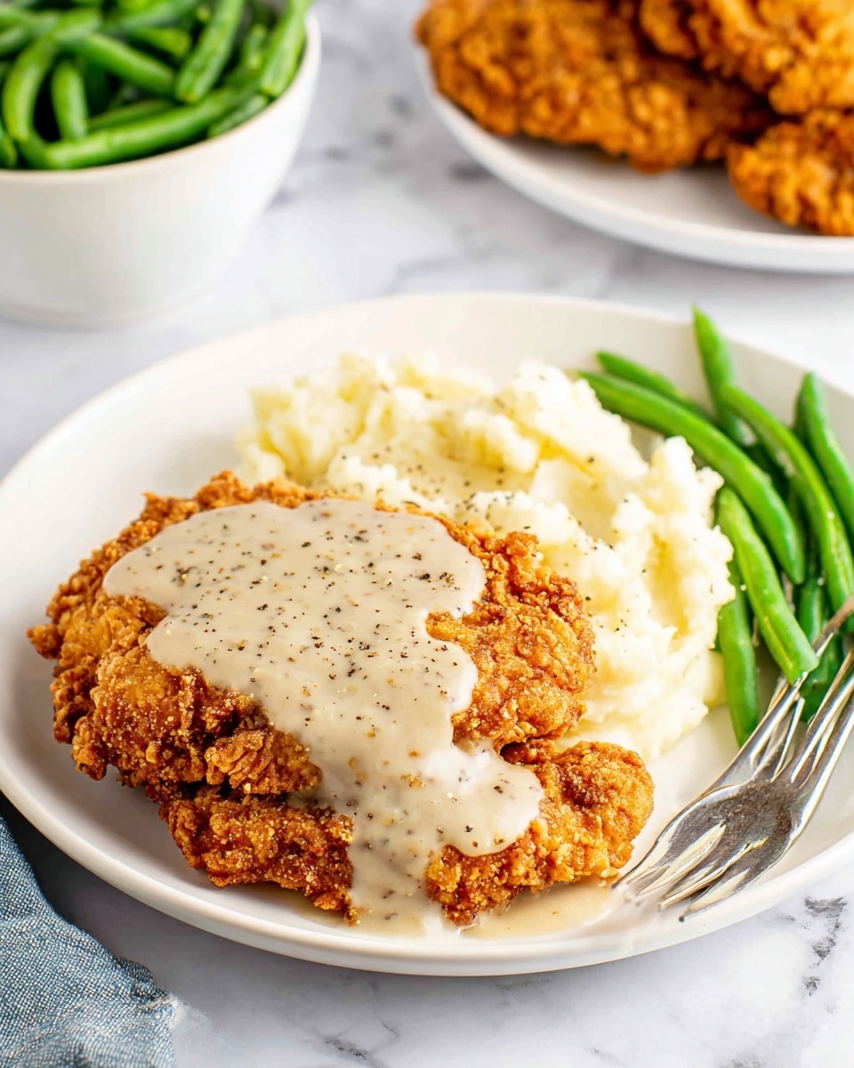 The image shows two breaded pork chops on a white plate with creamy sauce drizzled over the top of the front chop. The front pork chop is broken in half, showing a juicy, light gray inside with a crispy golden-brown crust. The sauce is light beige and creamy, with a few small green herb bits sprinkled on top. There are chopped green herbs scattered around the plate. A fork with a piece of pork chop covered in sauce is at the front left edge of the plate. The whole scene sits on a white marbled surface. photo taken with an iphone --ar 4:5 --v 7