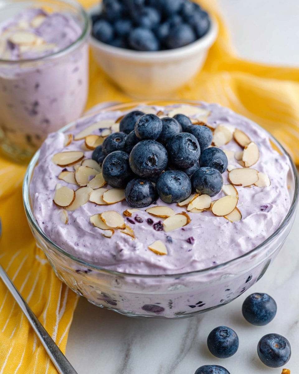 A close-up image shows a spoon holding a fluffy, light purple mixture with visible blueberries and a thin sliced almond on top. Below, a white bowl is filled with the same purple mixture that has a creamy texture with dark purple blueberry chunks inside. In the background, there is a larger white bowl also filled with the purple mixture, decorated with sliced almonds on top, and behind it, a bright yellow cup filled with fresh whole blueberries. A yellow striped cloth lies underneath everything on a white marbled surface with a blurred white tile wall behind. Photo taken with an iphone --ar 4:5 --v 7