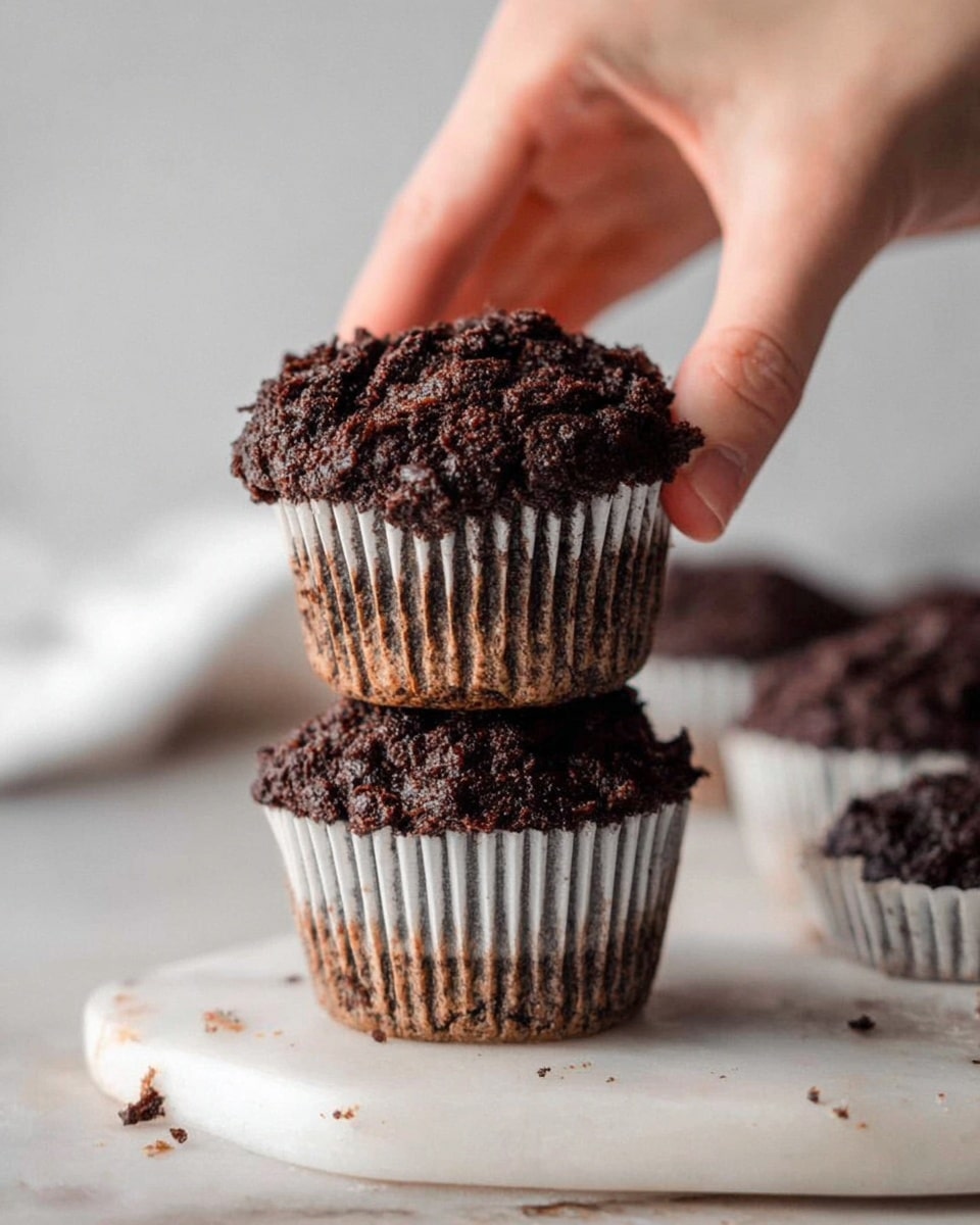 Two dark brown chocolate muffins with rough, crumbly tops are stacked, with the top muffin being held gently by a woman's hand. Both muffins are in white paper liners that have some brown stains near the base. They sit on a white marbled board with a few crumbs scattered around. In the blurred background, there are more muffins also in white paper liners. The overall setting has a soft, natural light coming from the side. photo taken with an iphone --ar 4:5 --v 7