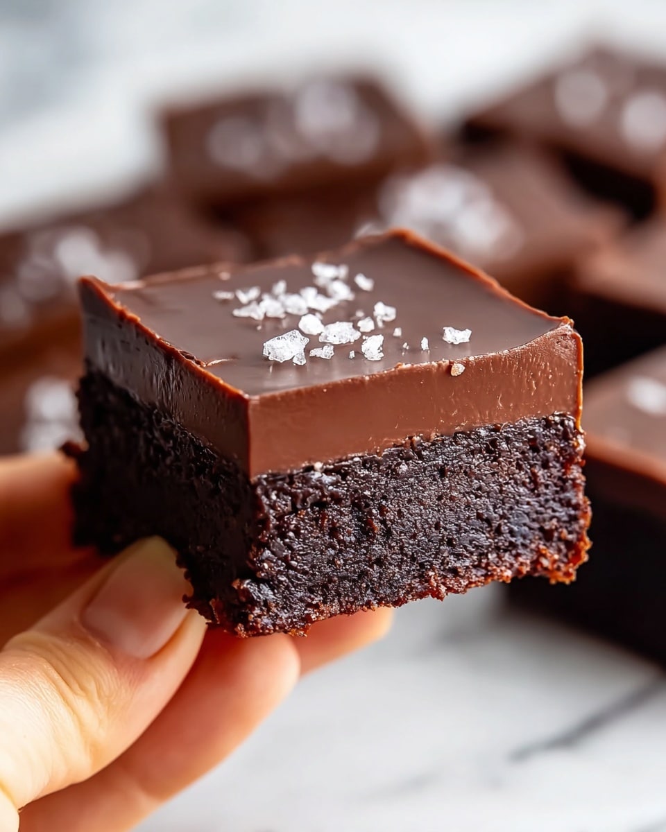 A close-up view of a two-layer chocolate dessert square held between a woman's hand thumb and finger shows a dense, dark chocolate brownie base with a rich, smooth milk chocolate ganache layer on top. The ganache is thick, glossy, and even, with a few small flakes of sea salt scattered on its surface. The edges of the dessert are clean-cut, and additional similar squares are blurred in the background on a white marbled texture. photo taken with an iphone --ar 4:5 --v 7