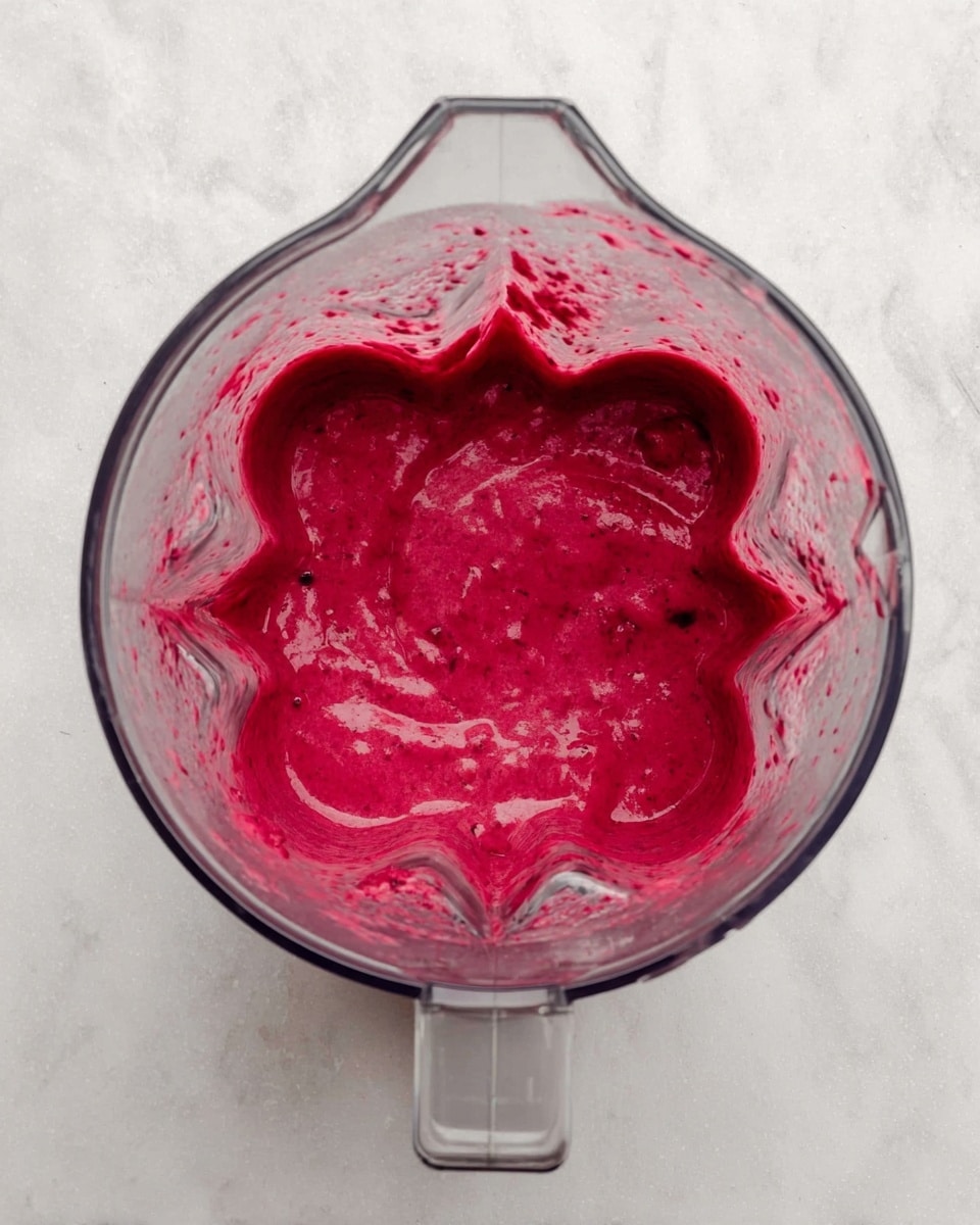 A glass jar filled to the top with a thick, bright red-purple smoothie with a frothy layer on top, a white straw inside the jar, a woman's hand pouring the smoothie from a clear blender jar. The jar sits on a white plate on a white marbled surface, with another glass jar of smoothie blurred in the background and half a dark red-purple beetroot on a white plate. Small sesame seeds are scattered near a small white plate filled with sesame seeds in the lower corner. photo taken with an iphone --ar 4:5 --v 7