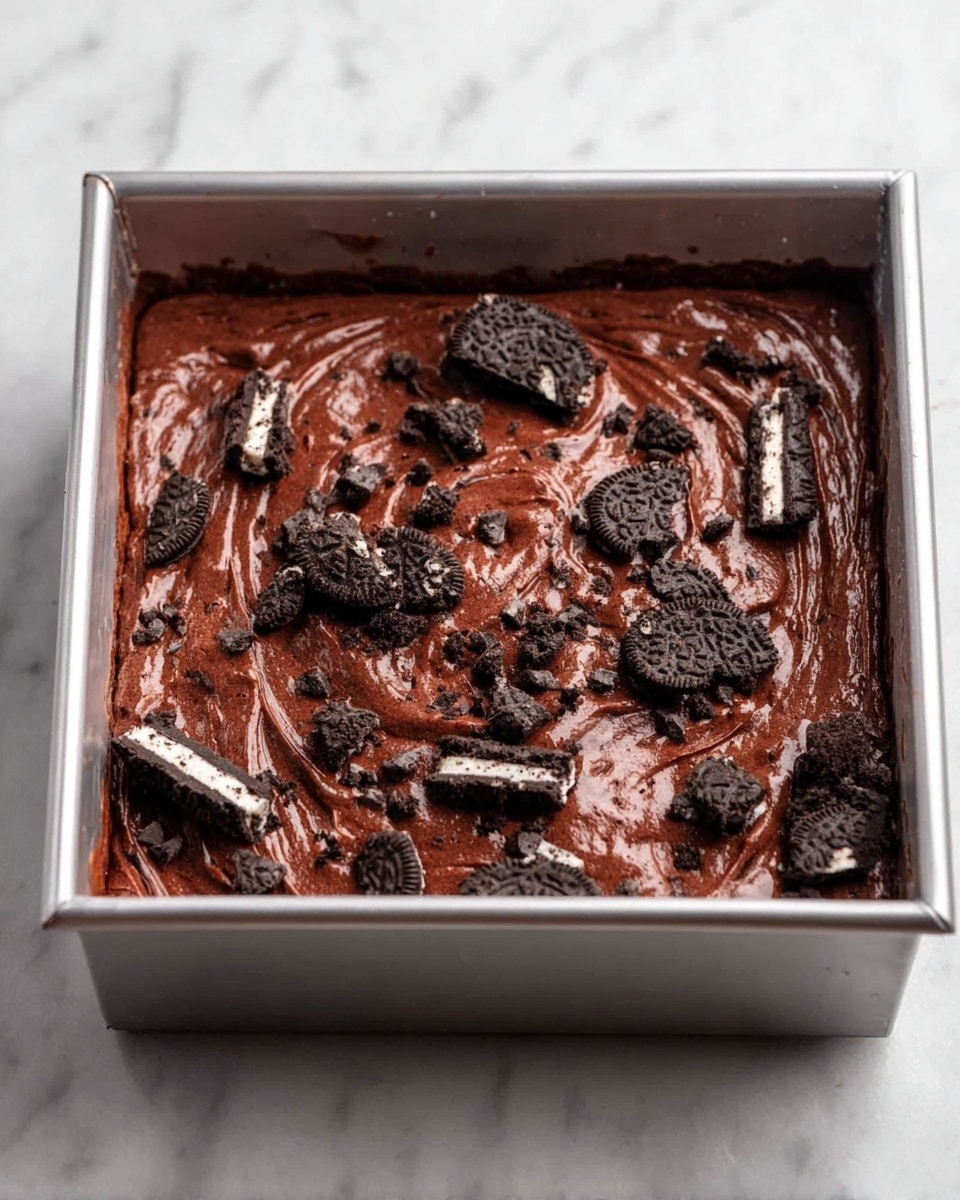 A square piece of rich, dark chocolate brownie sits on a white marbled surface, with a rough texture and moist inside. The brownie has two broken pieces of black and white cookies embedded on the top layer, showing the creamy filling and the dark cookie crumbs. In the background, blurred, is the larger remaining brownie with more cookie pieces on top and a visible missing section where the smaller piece was cut out. photo taken with an iphone --ar 4:5 --v 7