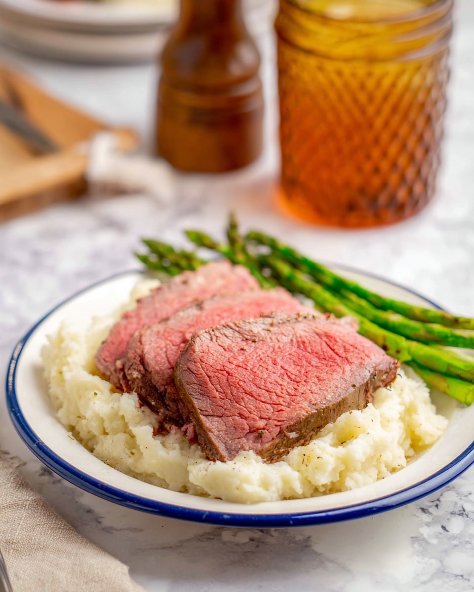 This image shows a white plate with a blue rim on a white marbled surface. On the plate, there is a base layer of creamy, white mashed potatoes with some texture showing small lumps. Above the mashed potatoes, there are two thick slices of medium-rare roast beef, with a pink center and browned outer edges. To the back side of the plate, there are several bright green asparagus spears arranged in a neat bundle. The background is softly blurred, featuring a pepper grinder and a glass jar with a textured amber liquid, along with a folded cloth napkin to the side. Photo taken with an iphone --ar 4:5 --v 7