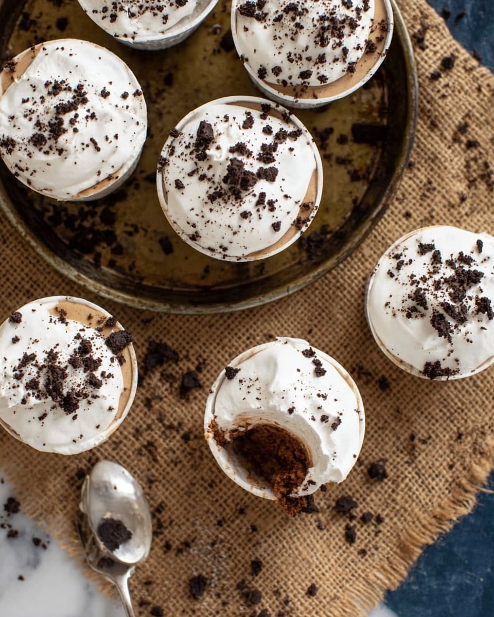 The image shows a small clear cup with three layers: the bottom layer is dark and crumbly, likely crushed cookies, the middle layer is smooth and light brown with small black specks, and the top layer is a thick dollop of white whipped cream sprinkled with dark cookie crumbs. More cups with the same dessert can be seen blurred in the background, placed on a rustic woven mat. The scene has a cozy feel with a white marbled texture underneath. photo taken with an iphone --ar 4:5 --v 7