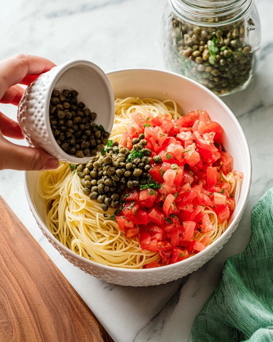 A white bowl holds a fresh pasta dish with spaghetti noodles as the base layer, pale yellow and smooth in texture. Mixed throughout are bright red, chopped tomatoes adding color and juiciness, along with scattered dark green capers that bring small pops of contrast. Fresh green parsley leaves are spread evenly across the dish, adding a lively touch on top. Zesty yellow lemon zest is sprinkled all over, brightening the appearance. The dish is placed on a white marbled surface, with two yellow lemons blurred in the background, enhancing the fresh and vibrant look. photo taken with an iphone --ar 4:5 --v 7